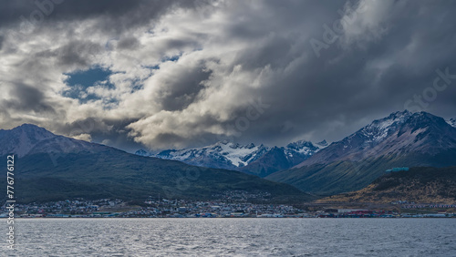 Beautiful snow-capped mountain range of the Andes against the sky and clouds. The city of Ushuaia is at the foot of the mountains, on the banks of the Beagle Canal. Ripples on the blue water.