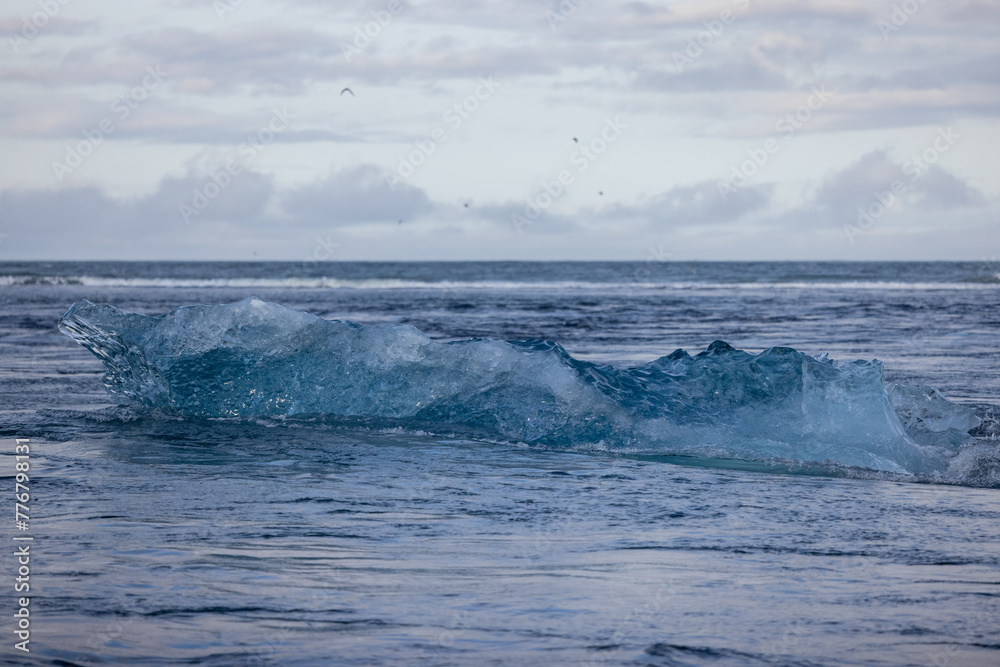 The powerful water wave crashes onto the sandy shore, creating a stunning natural landscape with the horizon blending into the cloudy sky. Jokulsarlon Glacier Lagoon, Iceland