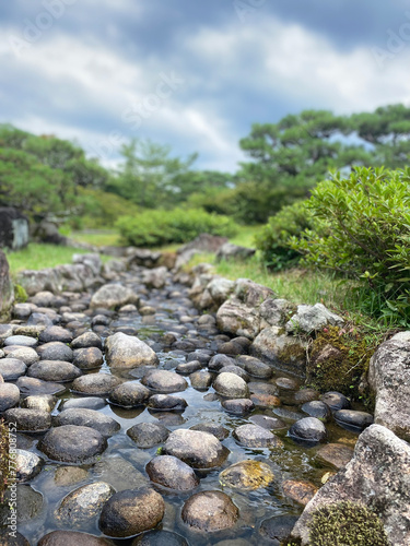 A tranquil pebble-filled stream bed with clear water flowing among the rounded stones. Lush greenery surrounds the stream under a partly cloudy sky.