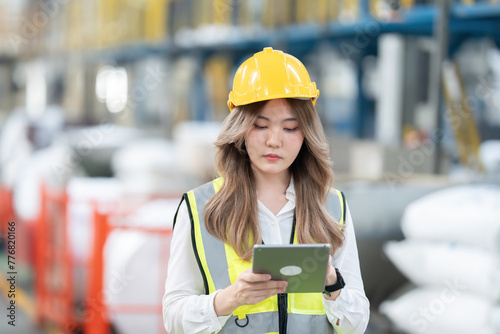 Long-haired Asian female engineer working intently looking at systems on laptop in industrial factory, business exporting products to international market of the world Wear a safety helmet and vest.
