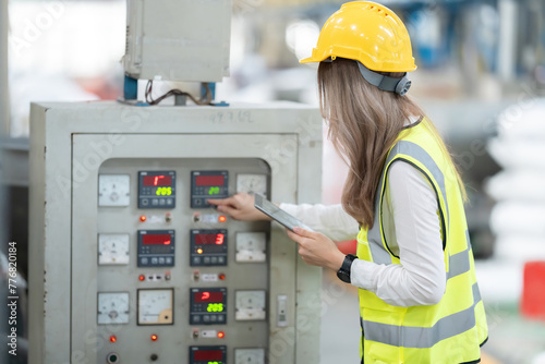 Asian female engineer with long hair pressing an electronic control panel controlling a system within an industrial factory, logistics business, holding a tablet. Wear a safety helmet and vest.