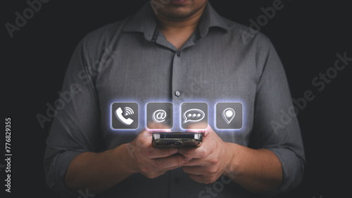 A man is holding a smartphone with contact icon include phone number, email, message and location, in contact us and marketing communication concept.