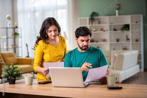 Young Indian couple checking mortgage or loan agreement, financial documents together, using laptop and calculator.
