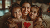 A woman is holding her two young daughters and a red heart-shaped box