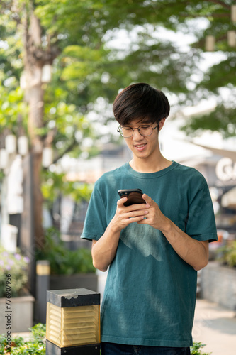 A young Asian man is using his smartphone, chatting with his friends while waiting them in a park.