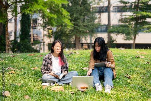 Two young Asian female college students are sitting on the grass in a park, using a laptop together.