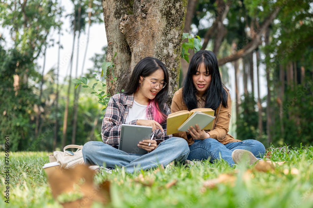 Two young Asian female students sit on the grass under a tree, studying together in a campus park.