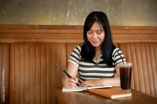 A young Asian woman is writing her diary or taking notes in her notebook at a coffee shop.