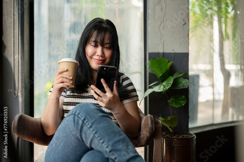 A happy Asian female is relaxing in a coffee shop, using her smartphone and enjoying her coffee.