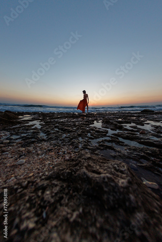 young woman in a summer dress walks along the seashore during sunset and looks at the sea.