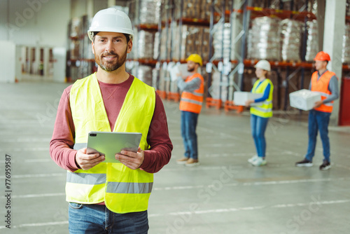 Bearded man, foreman wearing hard hat, holding digital tablet, supervises work of colleagues