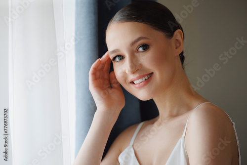 Happy young woman with makeup  radiant bride smiling softly by window. Concept morning and getting ready for wedding