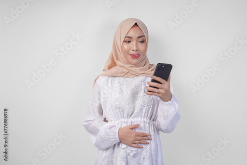 Portrait of young Asian Muslim woman in white dress and hijab feeling hungry while looking at her phone to order yummy food after fasting, isolated on white background