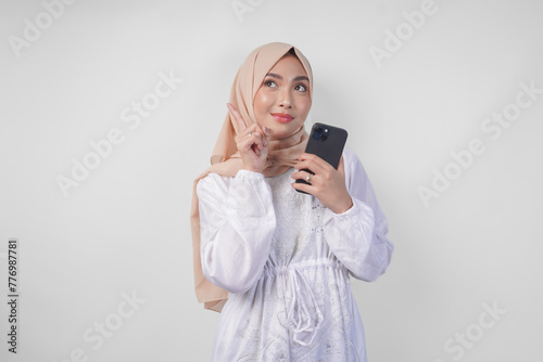 Thoughtful young Asian Muslim woman wearing white dress and hijab, using smartphone while holding her chin and thinking with serious expression over isolated white background