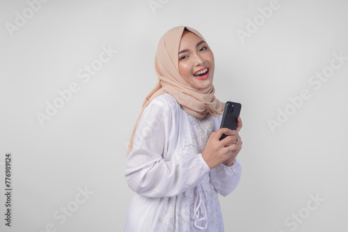 Excited Asian Muslim woman wearing white dress and hijab holding her smartphone while smiling cheerfully, isolated by white background. Ramadhan and Eid Mubarak concept
