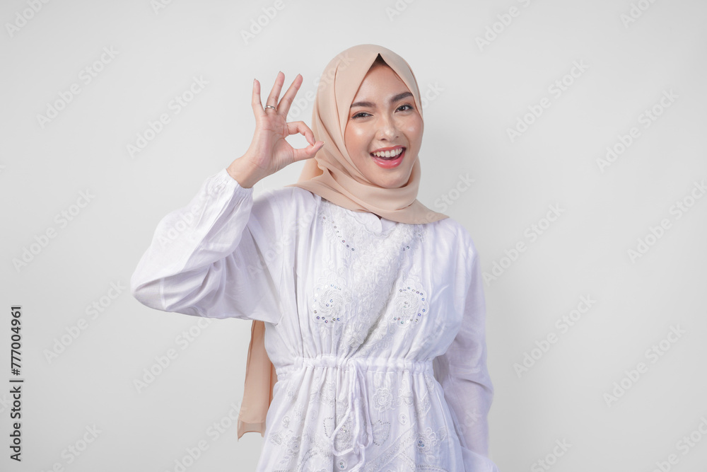 Fototapeta premium Portrait of Asian Muslim woman in white dress and hijab making an okay, good, nice sign gesture using fingers. Ramadan and Eid Mubarak concept
