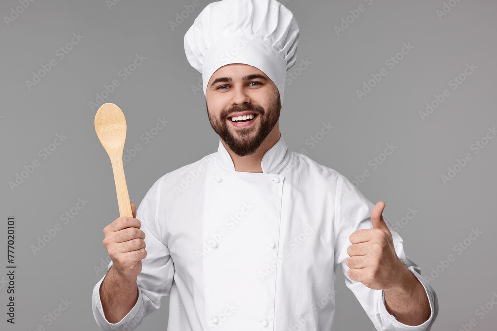 Happy young chef in uniform holding wooden spoon and showing thumb up on grey background Happy young chef in uniform holding wooden spoon and showing thumb up on grey background