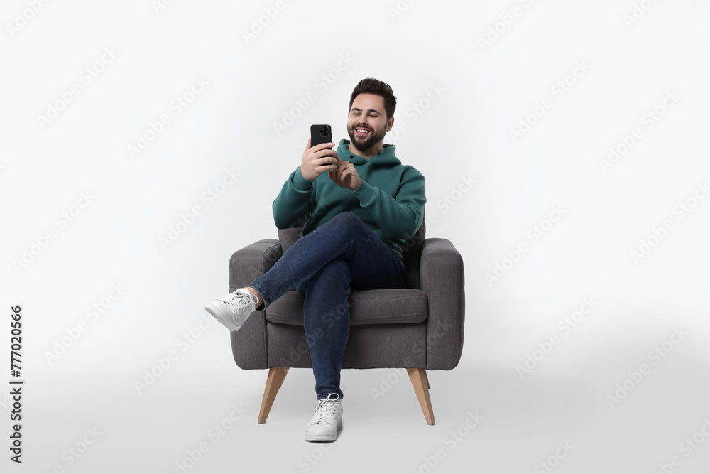 Happy young man using smartphone in armchair on white background