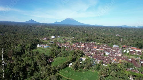 Aerial panorama of village surround with forest at centre of Bali island, camera fly back. Agung and smaller Batur mountains seen at background, numerous rice fields at large plain at south photo