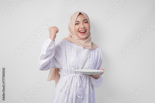 Cheerful young Asian Muslim woman in hijab doing a successful win gesture with clenched fist while presenting an empty plate and copy space over it © Reezky