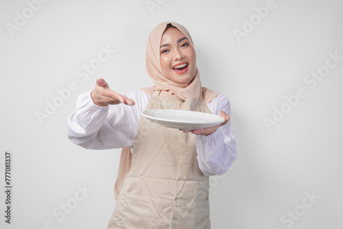 Portrait of a cheerful young Asian Muslim woman in a veil hijab and cream apron pointing to an empty plate with copy space over it, presenting food menu