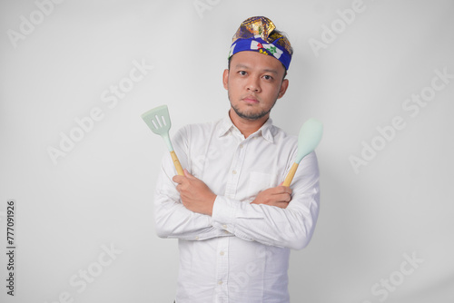 Young Balinese man in white shirt and traditional headdress holding spatula and kitchen cooking utensils with serious expression photo