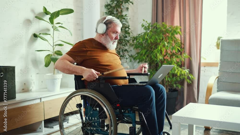 Elderly man sitting in wheelchair, wearing headphones and looking on laptop, learning how to play drums via online lessons. Home interior. Concept of healthcare, lifestyle, motivation, wellness, hobby Elderly man sitting in wheelchair, wearing headphones and looking on laptop, learning how to play drums via online lessons. Home interior. Concept of healthcare, lifestyle, motivation, wellness, hobby