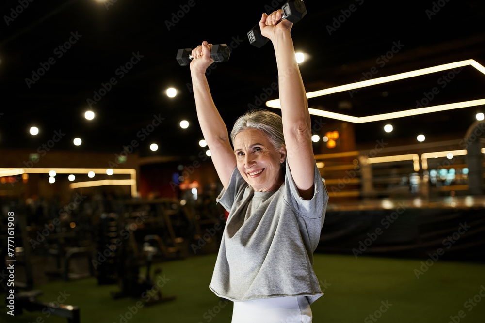 Fototapeta premium mature cheerful woman with gray hair in cozy attire training actively with dumbbells while in gym