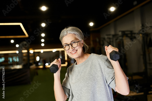 joyful mature woman with glasses and gray hair exercising with dumbbells and smiling at camera photo