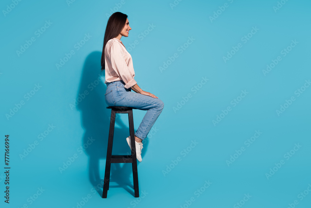 Full length photo of cute positive woman wear silky blouse sitting chair looking empty space isolated blue color background