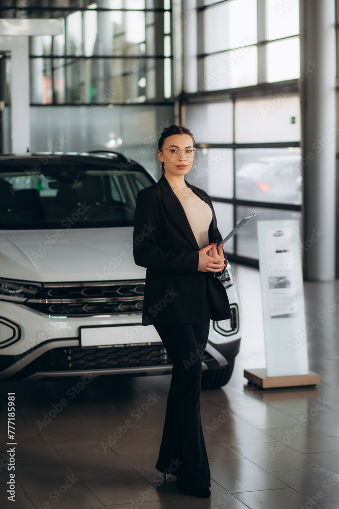 Smiling female seller in suit using tablet for looking which car is sold while standing in car salon. Smiling female seller in suit using tablet for looking which car is sold while standing in car salon.