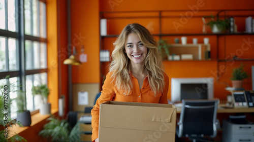 Young white woman, blonde, smiling, holding a box in an orange office.