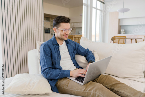 An affable man with a sincere smile works on a laptop, comfortably ensconced on a sofa in a light-filled, modern apartment.