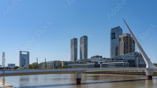 A beautiful pedestrian cable-stayed bridge Puente de la Mujer over the river. Stele and stretched metal strings against skyscrapers and city buildings. Clear blue sky. Argentina. Buenos Aires.