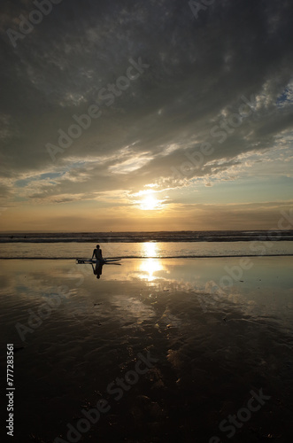 A man sitting on a surfboard on a sandy beach looking at the sunet ahead. Some dark clouds is rolling in and block some of the sunset. © Sophie