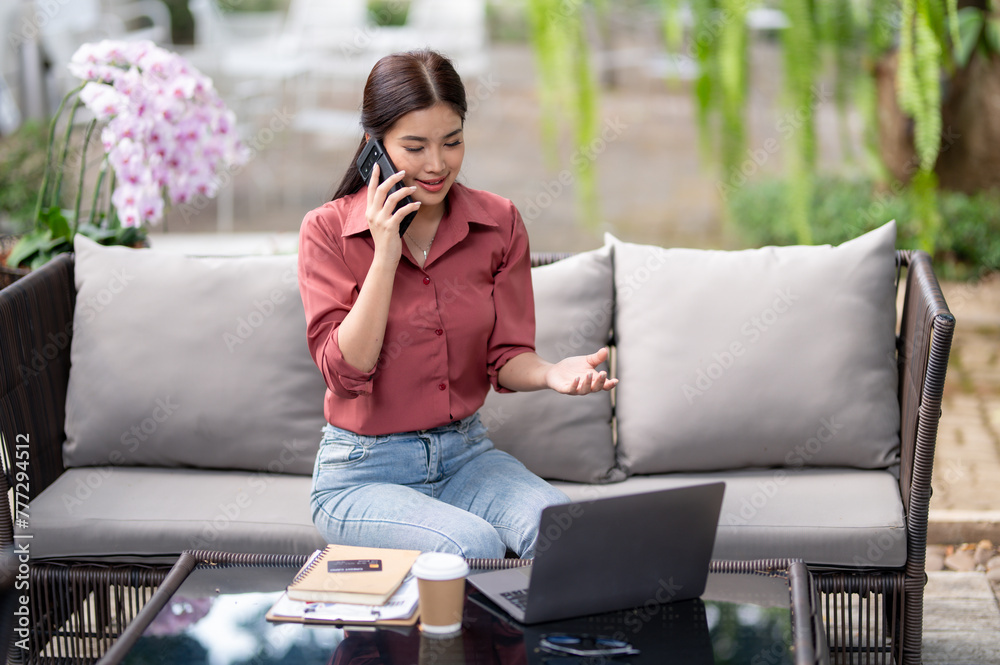 A businesswoman engages in a phone conversation while working on her laptop in a lush outdoor setting, showcasing multitasking. A businesswoman engages in a phone conversation while working on her laptop in a lush outdoor setting, showcasing multitasking.