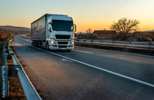 Big white truck driving on the highway through forested landscape at sunset. photo