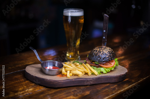 A burger with a knife sticking out of it and a glass of beer. The burger is black and has a lot of toppings. The fries are on a plate next to the burger