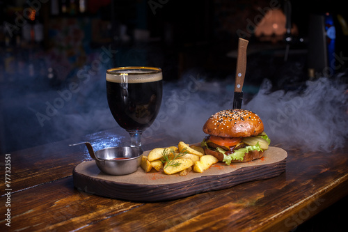 A burger and fries are on a wooden table with a glass of beer. The burger is topped with lettuce and tomato, and the fries are seasoned.