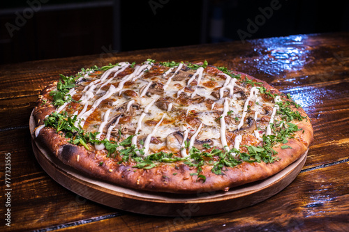 A pizza with cheese and herbs sits on a wooden table. The pizza is topped with a green herb garnish and has a creamy sauce