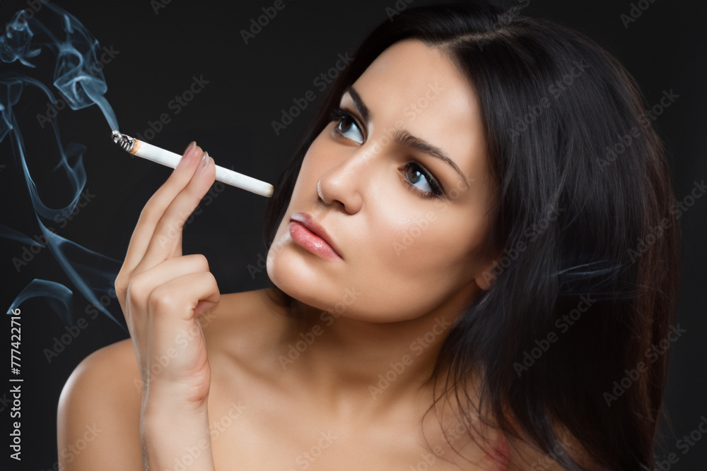 Fototapeta premium girl with a cigarette. young sexy girl with black long hair sits with bare shoulders on a black background with a cigarette in her fingers, harm concept
