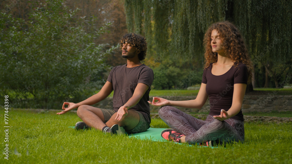 Two diverse multiracial guy and girl meditating zen meditation morning sport practice in park lotus position on yoga mat on grass sporty Indian Arabian man Caucasian woman relaxing in nature outdoors Two diverse multiracial guy and girl meditating zen meditation morning sport practice in park lotus position on yoga mat on grass sporty Indian Arabian man Caucasian woman relaxing in nature outdoors