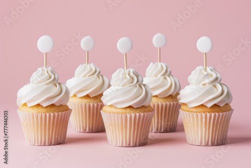 Five vanilla cupcakes with fluffy white frosting and blank circular toppers ready for customization, on a pink background.