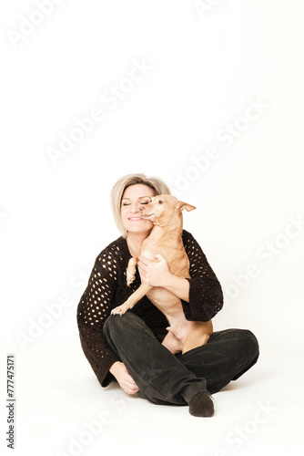 Blonde woman dessed in black clothes hugging her brown hound seated in a white background