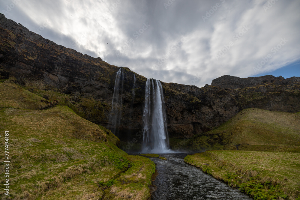 Fototapeta premium Skogafoss waterfall cascades down surrounded by mountains and green grass on a cloudy day, creating a picturesque natural landscape of fluvial landforms. Iceland