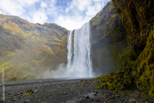 A stunning Skogafoss waterfall cascades down a mountain chute in a valley, surrounded by towering mountains under a clear blue sky with fluffy clouds. Iceland
