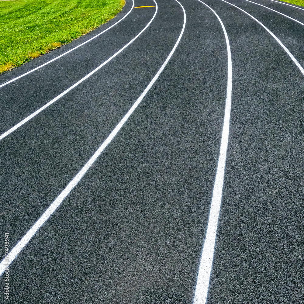 Track and Field Running Lanes. Overhead view of a rubber black running track surface with slightly curved white lane lines. Natural grass surface on the inside of the circular track.