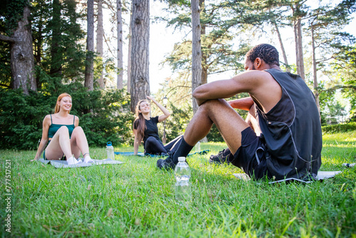 Three people are sitting on the grass in a park during a workout together on a summer day