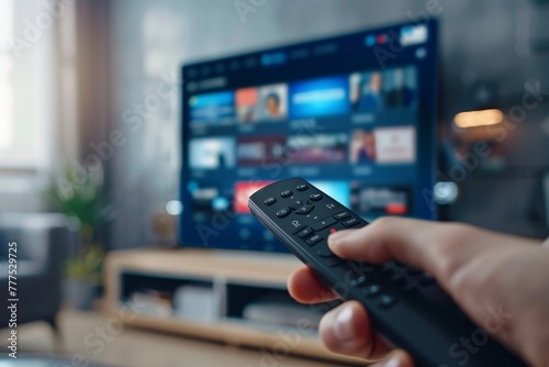 A hand holding a remote control in front of a smart TV with streaming services on the screen, in a close up shot.