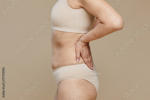 Side view of aged female model wearing beige brasserie and panties touching her back during photo session while posing in isolation photo